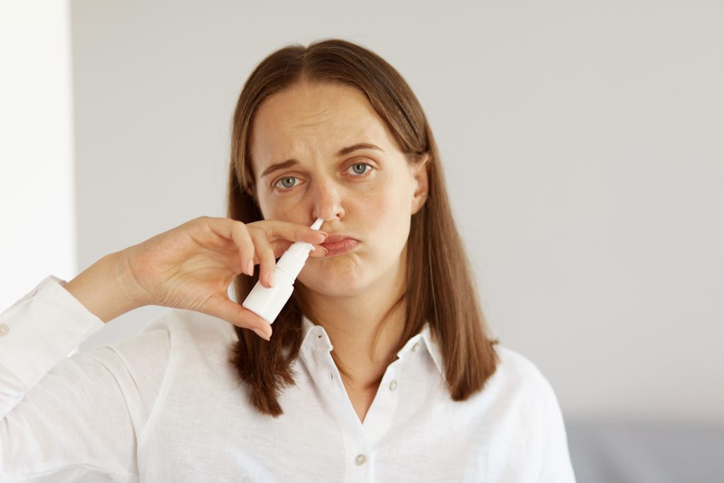 Темноволосая женщина в белой рубашке брызгает сосудосуживающий спрей в нос