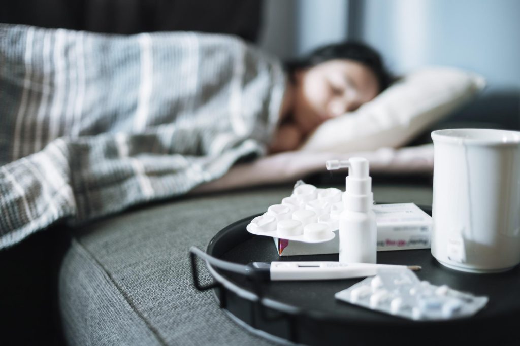 На столе стоят лекарства от насморка, в кровати на фоне лежит девушка